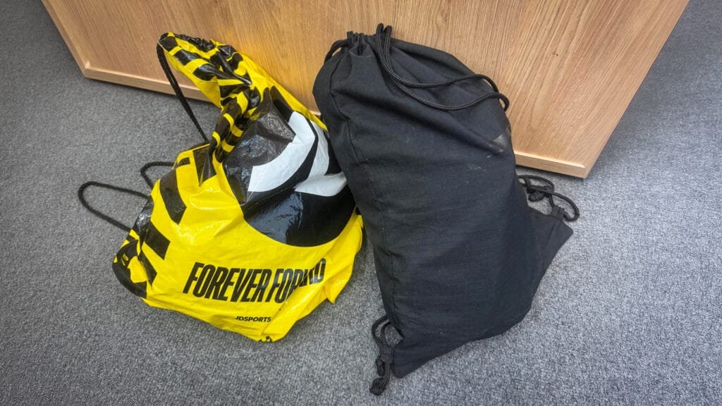 A yellow JD Sports drawstring bag with bold black text reading ‘FOREVER FORWARD’ next to a black drawstring bag, both placed on a grey carpet in front of a wooden cabinet.