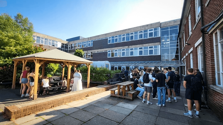 A group of students gathered in a school courtyard with brick buildings and large windows, standing near wooden benches and a covered seating area in bright sunlight.