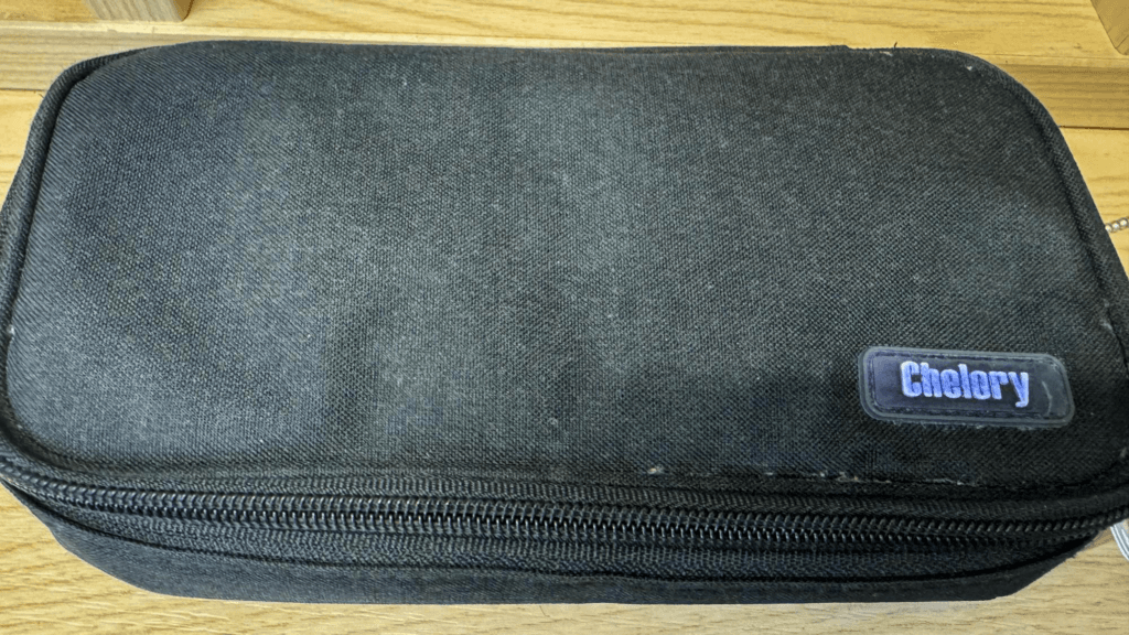 A black fabric pencil case with a zip fastening and a small ‘Chelory’ brand label, placed on a wooden surface.