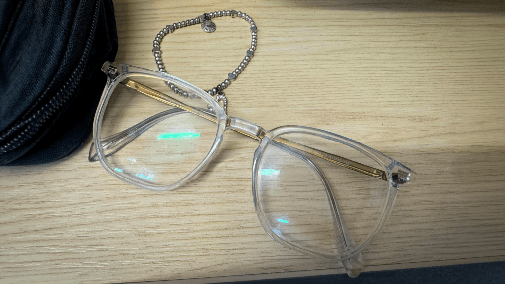 A pair of clear-framed glasses resting on a wooden desk beside a black case and a silver beaded bracelet with small charm details.
