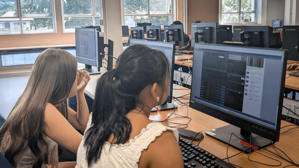 Students working at desktop computers in a computer suite, viewing software on large monitors, with rows of workstations and windows in the background.