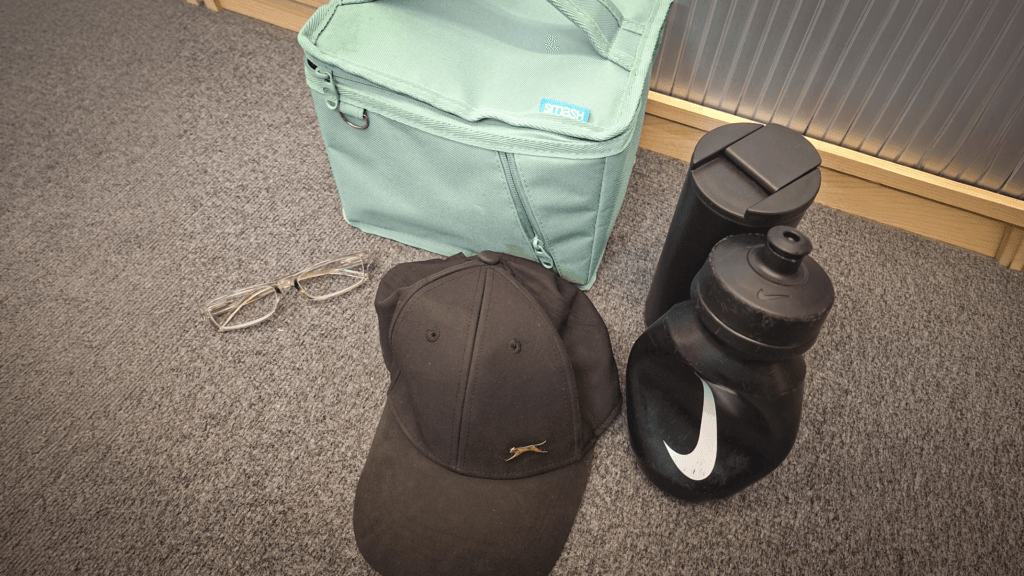 A mint green insulated lunch bag, clear-framed glasses, a black baseball cap, a black reusable drinks bottle, and a black travel mug arranged on a grey carpet.