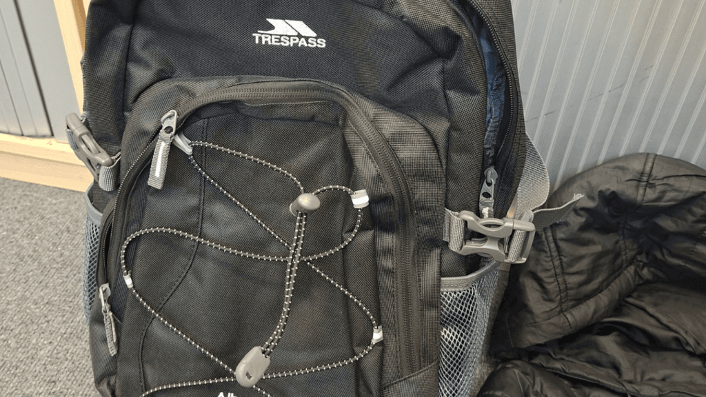 Close-up of a black Trespass backpack with several zipped compartments and elasticated front cords.