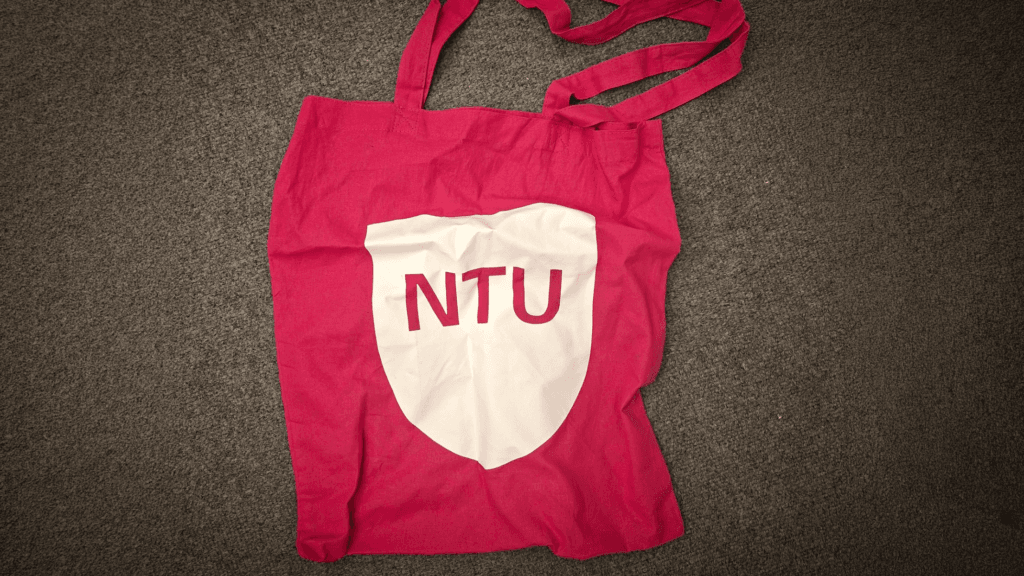 A pink fabric tote bag featuring a white shield design with the letters ‘NTU’ printed in the centre, laid flat on a grey carpet.