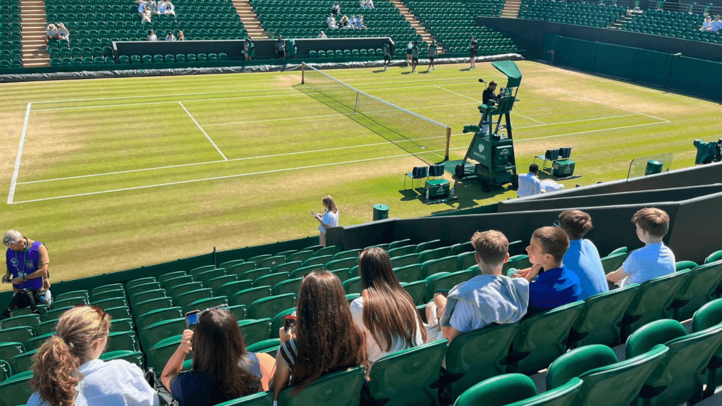Students seated in the stands overlooking a grass tennis court at Wimbledon.