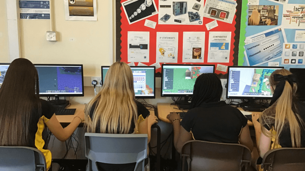 Four students using desktop computers in an ICT classroom. The monitors display Minecraft gameplay, and educational posters covering computing topics such as binary numbers, algorithms, login credentials, and programming concepts are displayed on the wall behind them.