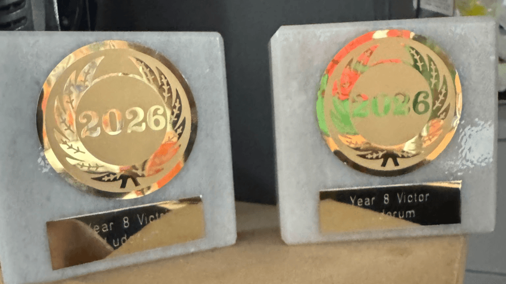 Two small award trophies displayed side by side on a shelf. Each trophy features a gold circular emblem with “2026” in the centre, surrounded by a laurel wreath design, mounted on a light grey rectangular base. A black nameplate on each trophy includes the words “Year 8 Victor” and “Judicum.”