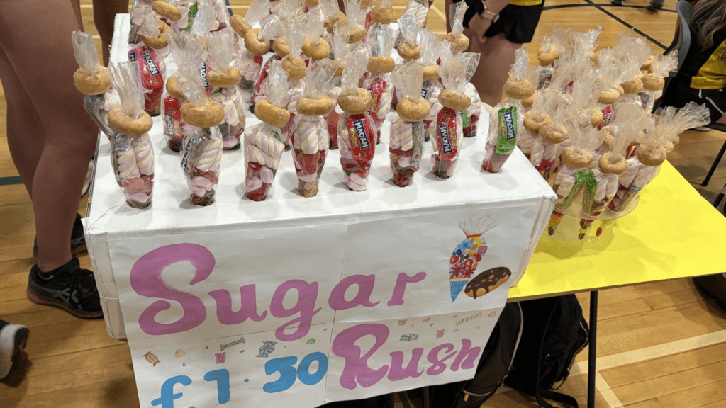 Fundraising stall displaying numerous cellophane-wrapped sweet cones filled with marshmallows and assorted confectionery. A large handmade sign on the front reads “Sugar Rush £1.50.” The stall is set up in a sports hall with people standing nearby.