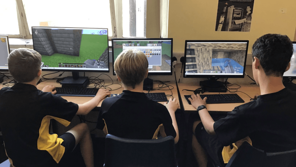 Three students seated at desktop computers in a classroom, each playing Minecraft on separate monitors. The screens display building and construction activities within the game, while keyboards and mice are arranged on a desk beneath large windows.