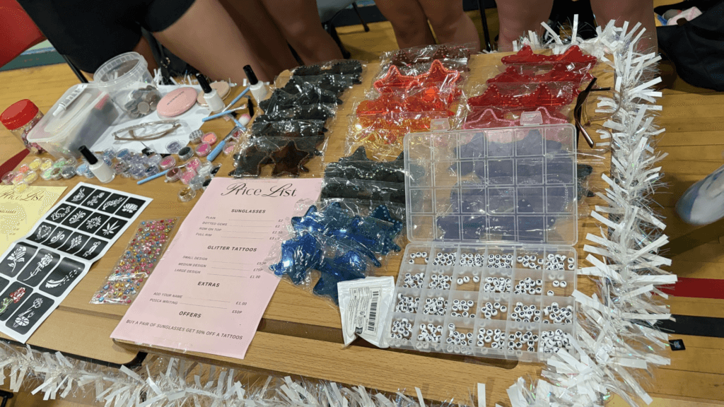 Craft and accessories stall displaying colourful star-shaped sunglasses, temporary tattoo stencils, decorative gems, letter beads for custom jewellery, and a printed price list. White tinsel decorates the table, which is located in an indoor hall during a fair or market event.