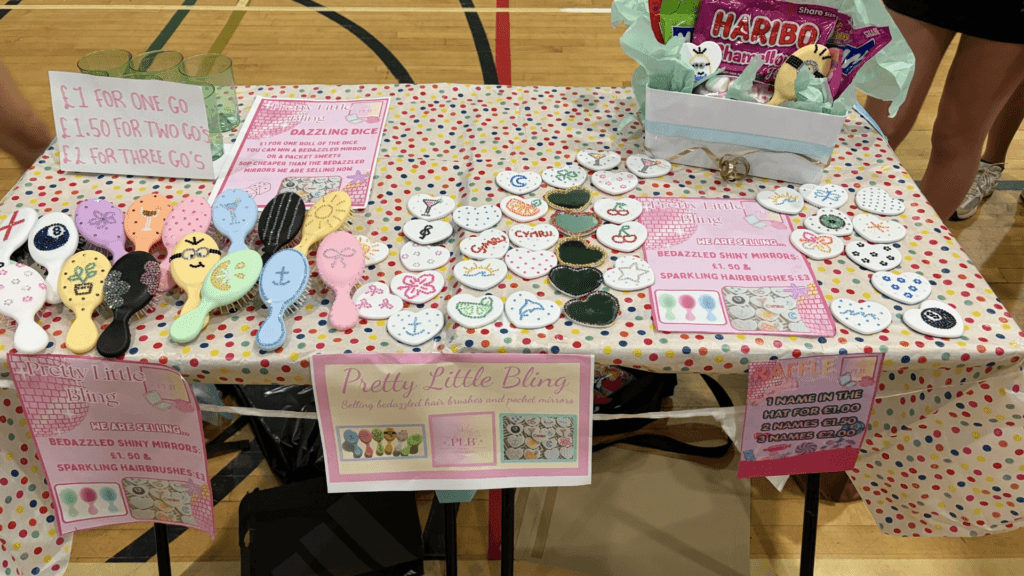 Table display at a school or community fair featuring decorated handheld mirrors and hairbrushes with colourful bead designs. Signs advertise “Pretty Little Bling” products including bedazzled mirrors, sparkling hairbrushes, a raffle, and a dice game. The table is covered with a multicoloured polka-dot cloth and includes a gift basket of sweets.