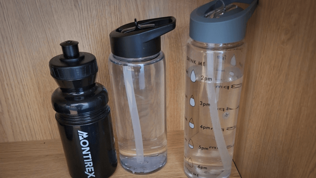 Three reusable water bottles displayed on a wooden shelf: a black Montirex sports bottle, a clear bottle with a black lid and built-in straw, and a larger clear hydration bottle with a grey lid, straw, and hourly drink-tracking markings.