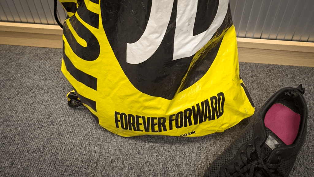 Yellow and black sports bag with the slogan “FOREVER FORWARD” printed on the front, placed on a grey carpet. Part of a black football boot with a pink insole is visible beside the bag.