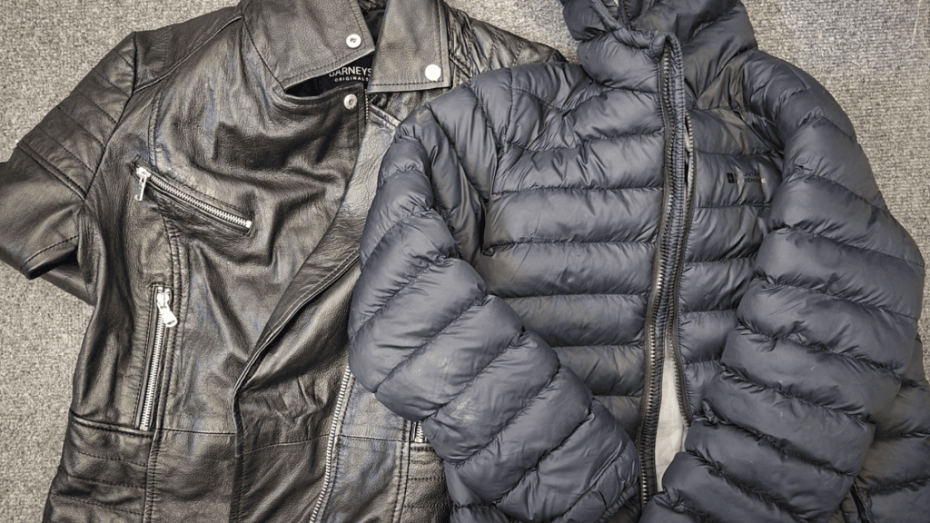 Two black outerwear garments laid flat on a grey carpet: a black leather-style biker jacket with silver zip details and a black quilted puffer jacket with a hood and front zip fastening.