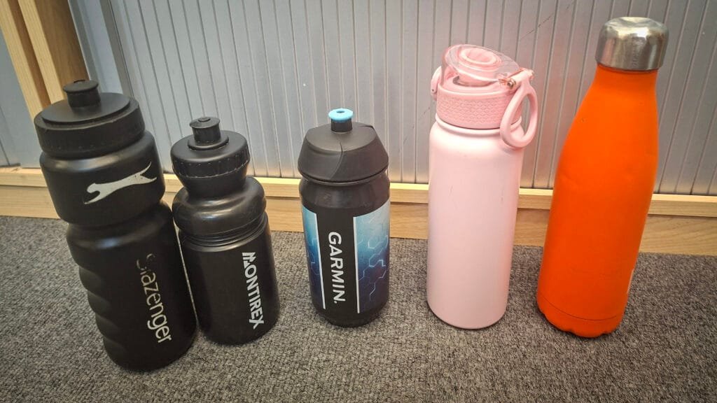 Five reusable water bottles displayed on a grey carpet against a panelled wall, including two black sports bottles, a black Garmin-branded bottle with blue detailing, a pale pink insulated bottle with a carry handle, and a bright orange insulated metal bottle.