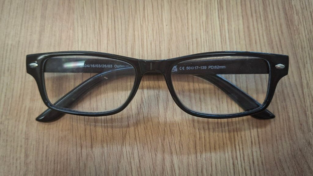 Pair of black rectangular glasses resting on a wooden surface. The frames have clear lenses and small silver-coloured rivet details at the outer corners.