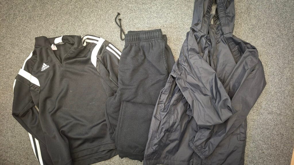 Black sports clothing laid out on a grey carpet, including an Adidas long-sleeved training top with white stripes, black joggers, and a black hooded waterproof jacket.