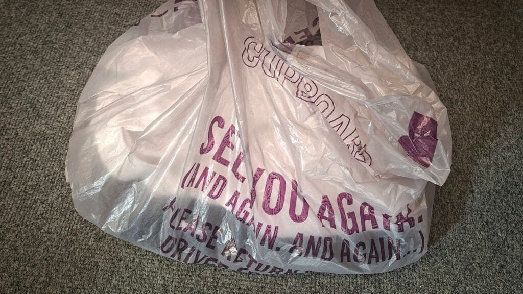 White plastic carrier bag resting on a grey carpet. Purple text on the bag includes the words “See you again and again and again” and “Please return.”