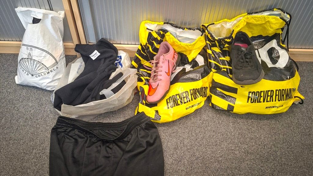 Assortment of clothing and footwear arranged on a grey carpet, including black sports shorts, dark clothing in carrier bags, two yellow and black JD Sports bags with the slogan “FOREVER FORWARD,” a pink football boot, a black trainer, and a white shopping bag featuring a hot-air balloon illustration and the text “Tenerife Columbina.”
