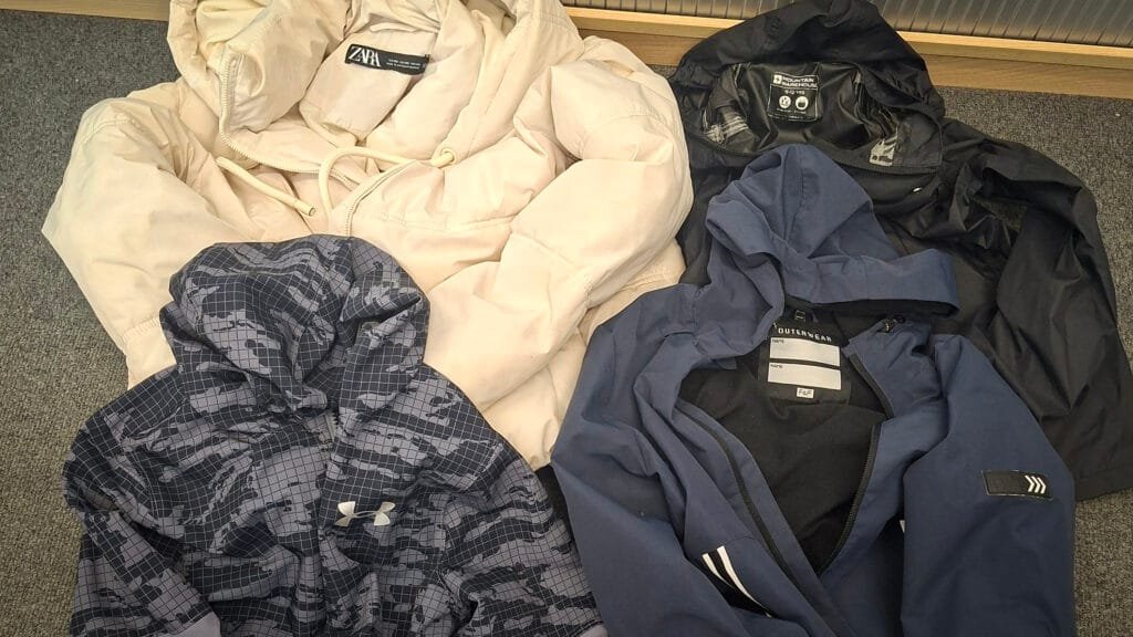 Collection of outerwear laid out on a grey carpet, including a cream Zara puffer jacket, a black hooded waterproof jacket, a blue hooded jacket, and a blue and grey patterned Under Armour zip-up sports hoodie.