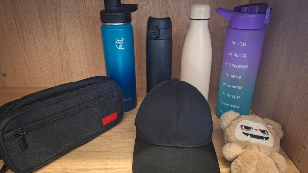 Items displayed on a wooden shelf, including four reusable water bottles in blue, black, cream, and purple, a black pencil case, a black baseball cap, and a small beige plush toy with a cartoon-style face. The purple bottle features printed hydration-time reminders.