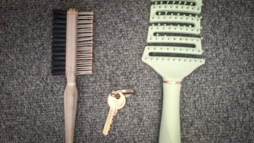 Haircare items placed on a grey carpet, including a mint-green vented hairbrush, a small double-sided brush with bristles and metal pins, and a single silver-coloured key attached to a keyring.