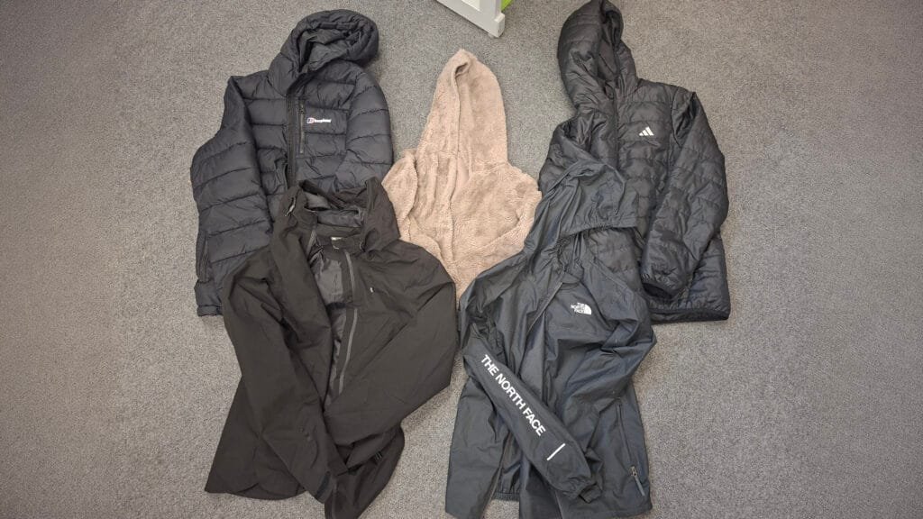 Collection of coats and jackets laid out on a grey carpet, including a black Berghaus hooded puffer jacket, a beige fleece or dressing gown-style garment, a black Adidas hooded puffer jacket, a black waterproof jacket, and a dark grey The North Face waterproof jacket.