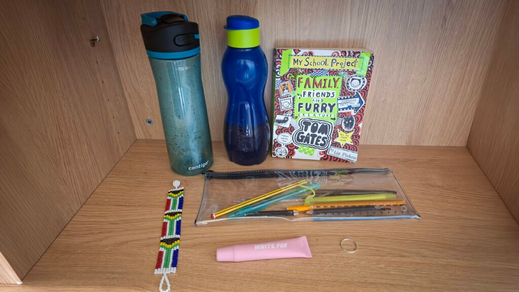 Personal and school items displayed on a wooden shelf, including two reusable water bottles, a book titled “My School Project: Family, Friends and Furry Creatures” by Tom Gates, a clear pencil case containing stationery, a colourful beaded bracelet, a pink tube of lip balm, and a silver-coloured ring.