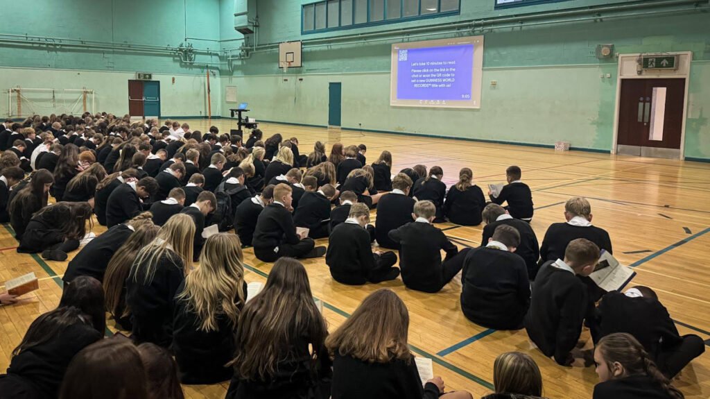 Large group of students seated on the floor of a sports hall taking part in a whole-school reading activity. A presentation screen at the front displays instructions encouraging participants to spend ten minutes reading and contribute towards a reading challenge or world record attempt.