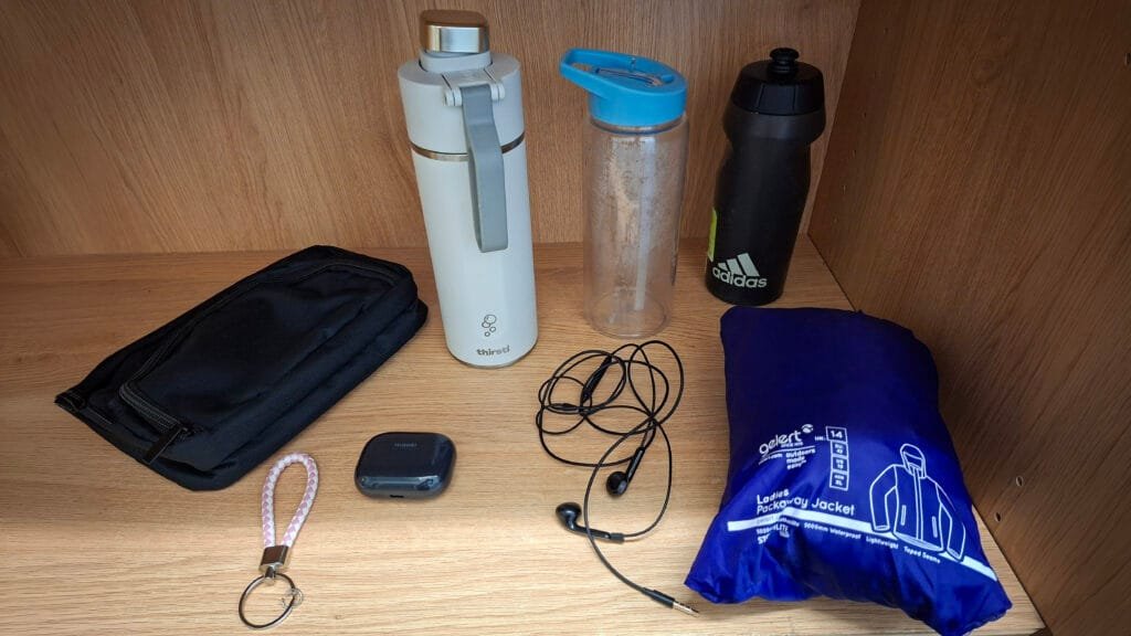 Items arranged on a wooden shelf, including a black pencil case, three reusable water bottles, a Huawei wireless earbud charging case, wired earphones, a pink-and-white wrist strap with key ring, and a blue packaway waterproof jacket stored in its matching pouch.