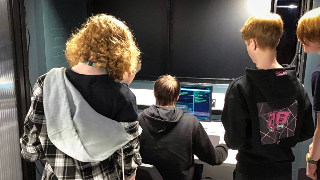Students gathered around a computer workstation in a technology or cybersecurity classroom. One student is seated using a computer displaying code or system-monitoring software, while other students stand nearby observing the screen and discussing the activity. The room has dark walls and specialist computing equipment.