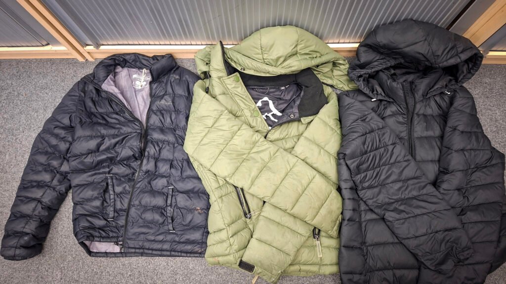 Three quilted hooded puffer jackets laid out on a grey carpet: a black Mountain Warehouse jacket, a light green puffer jacket, and a black puffer jacket with a hood. All three coats are displayed flat for viewing.