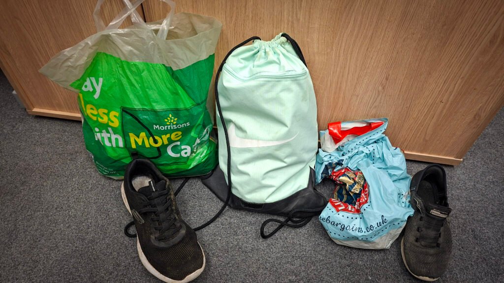 Collection of personal belongings on a grey carpet, including a green Morrisons carrier bag, a mint-green Nike drawstring bag, a blue shopping bag, and a pair of black trainers positioned on either side of the bags.