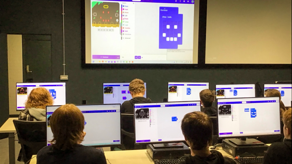 Students seated at desktop computers in an ICT classroom learning to program BBC micro:bit devices using the Microsoft MakeCode interface. A large presentation screen at the front of the room displays coding blocks and an LED smiley-face pattern while students follow along on their individual monitors.