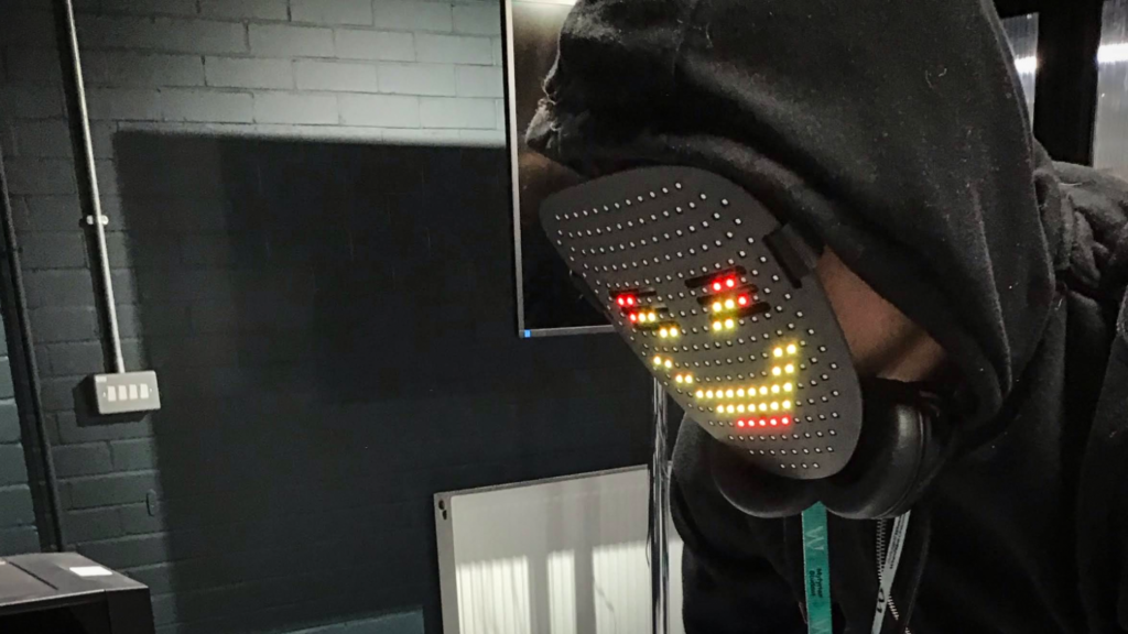 Student wearing a hooded top and headphones while demonstrating a wearable LED face mask displaying a pixelated smiling face using red, yellow, and white lights. The activity appears to take place in a computing or technology classroom.