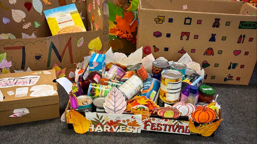 Harvest Festival display featuring decorated collection boxes and trays filled with donated food items, including tins, pasta, sauces, cereal, and other pantry essentials. Autumn-themed decorations such as leaves, pumpkins, and harvest artwork surround the donations.