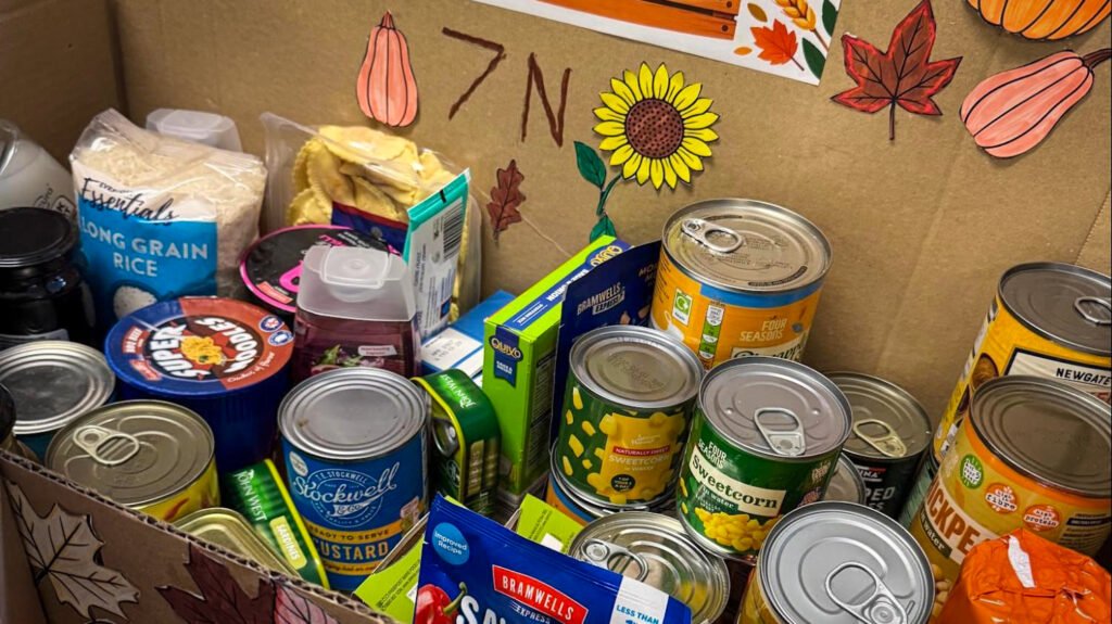 Close-up of a Harvest Festival food donation box containing a variety of non-perishable items, including canned vegetables, soup, rice, custard, pasta sauce, and other groceries. The display is decorated with sunflower, pumpkin, and autumn leaf artwork, with “7N” written on the background panel.