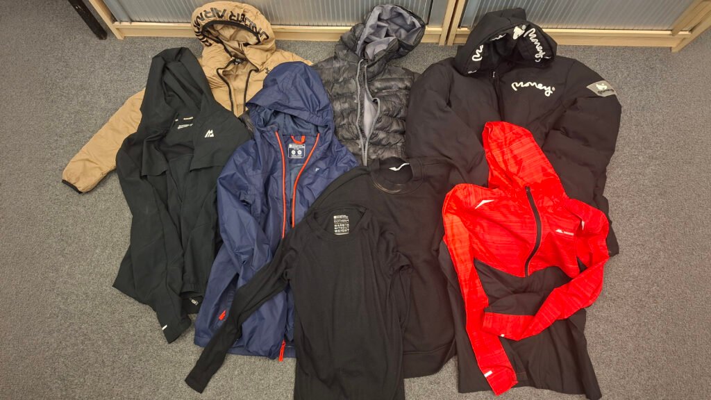Collection of clothing laid out on a grey carpet, including a tan hooded puffer jacket, a black waterproof jacket, a blue hooded rain jacket, a grey quilted jacket, a black puffer coat with “Money” branding, a black sweatshirt, and a bright red zip-up sports jacket.