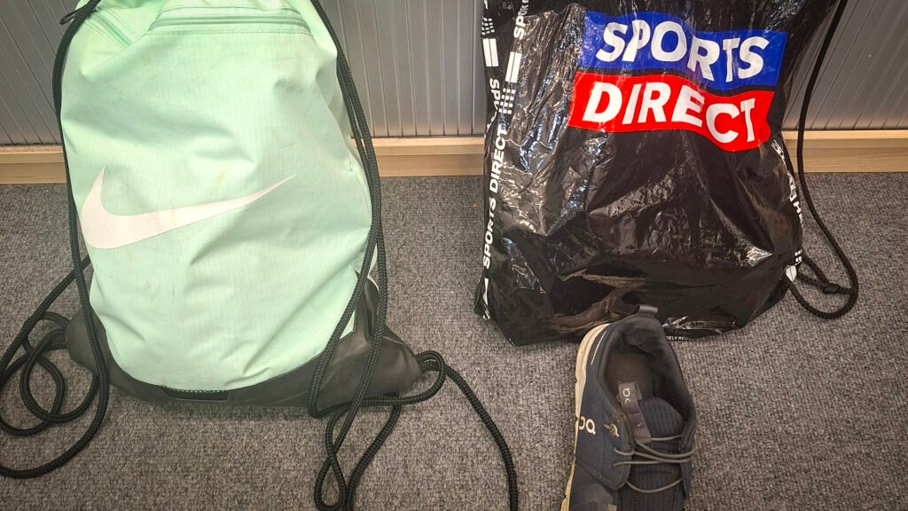 Personal belongings arranged on a grey carpet, including a mint-green Nike drawstring bag, a black Sports Direct carrier bag, and a single dark-coloured sports trainer positioned in front of the bags.