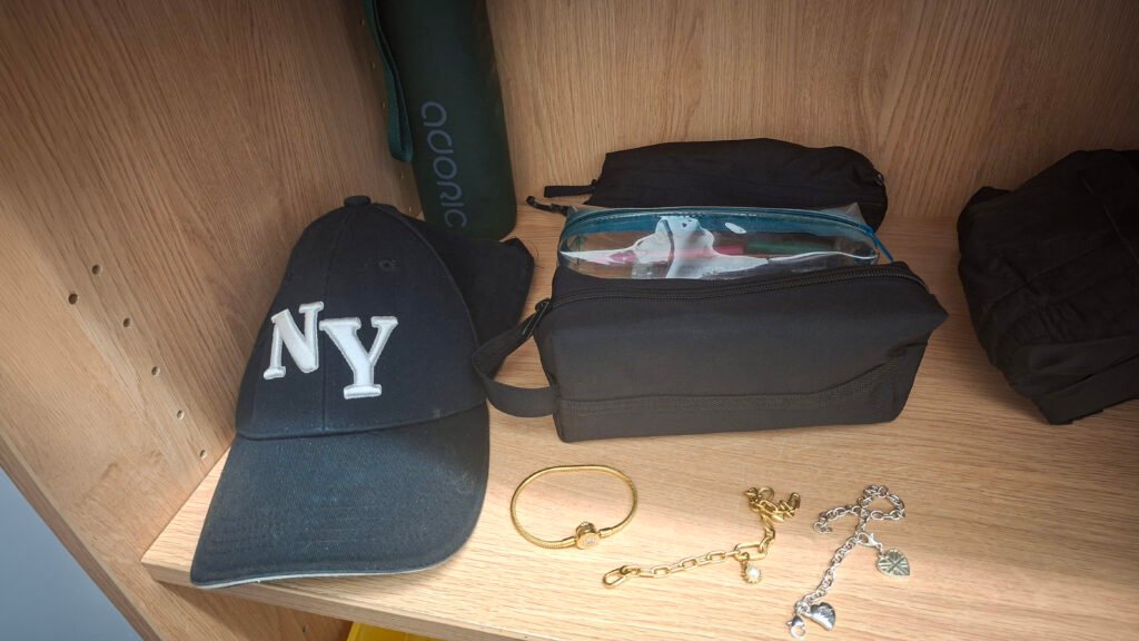 Items arranged on a wooden shelf, including a black New York Yankees baseball cap, a tall dark-green water bottle, two black pencil cases, a clear pencil case containing stationery, a gold-coloured bracelet, a gold-coloured chain bracelet with a charm, a silver-coloured chain bracelet with heart-shaped charms, and a folded black item of clothing. The shelf is set against a wooden cabinet backdrop.