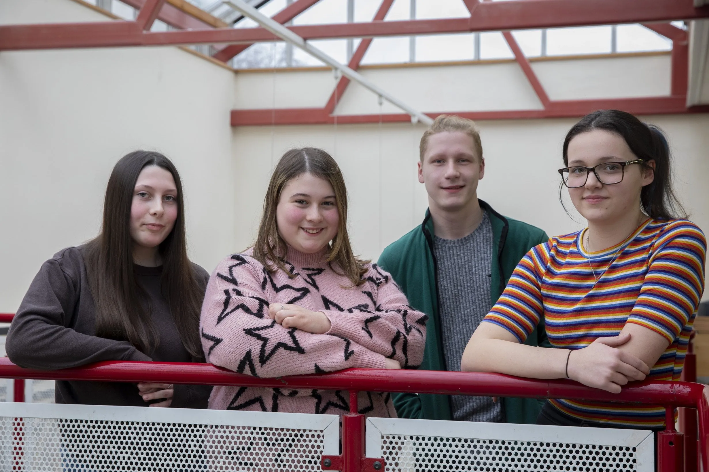 Four sixth form students standing together indoors, leaning on a red railing and looking towards the camera.