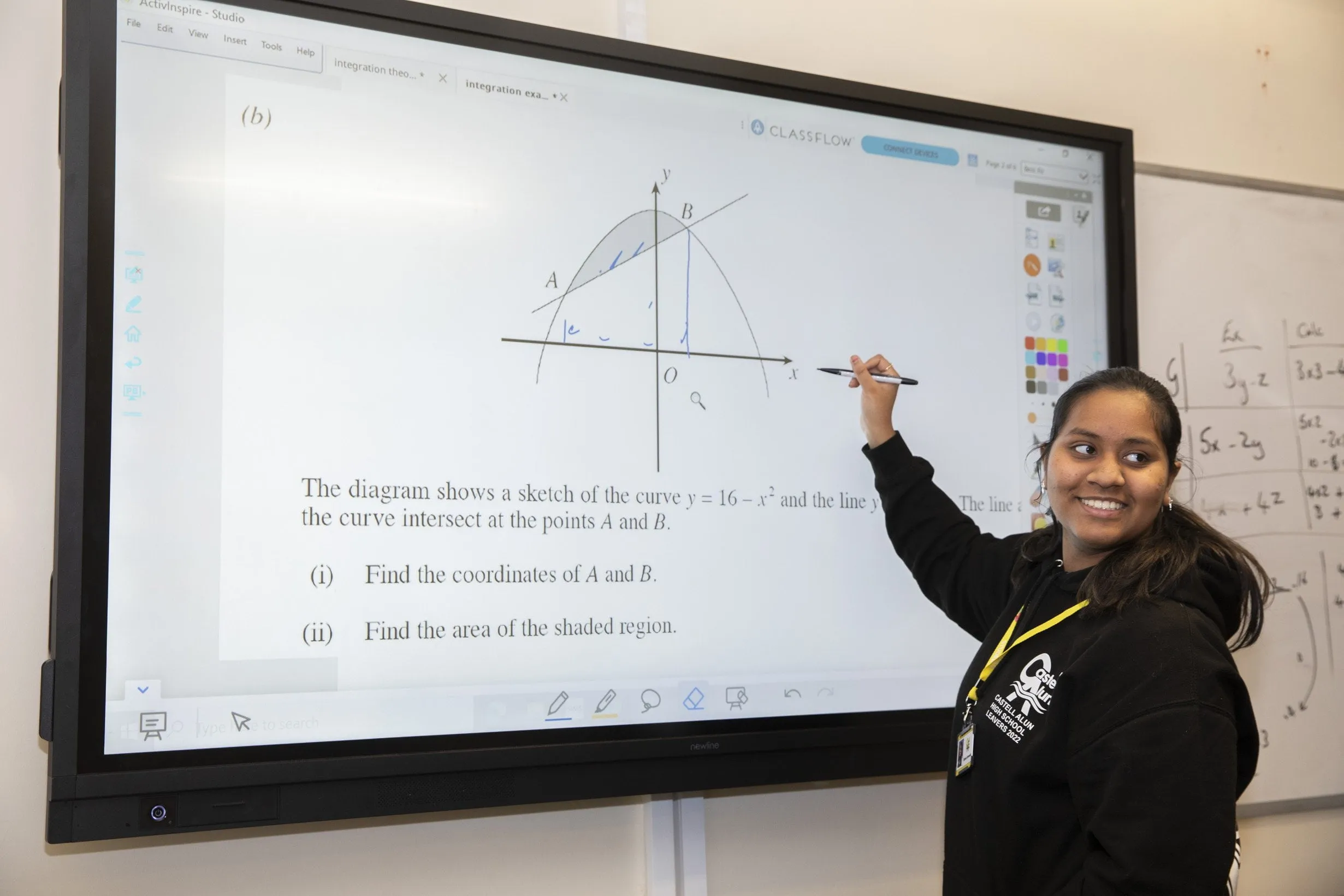 A student standing beside an interactive classroom display, pointing to a mathematics diagram showing a curve and a straight line, with mathematical questions and working visible on the screen.