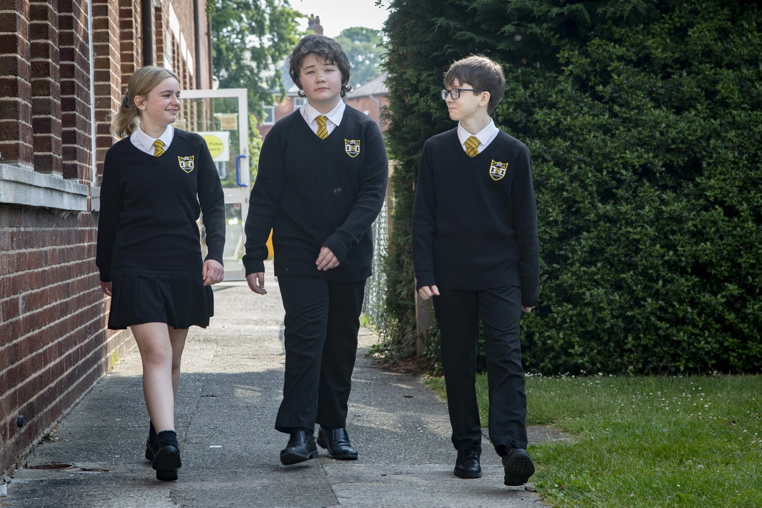 Three students in school uniform walking along a paved path beside a brick school building, with trees, hedges, and school grounds visible in the background.
