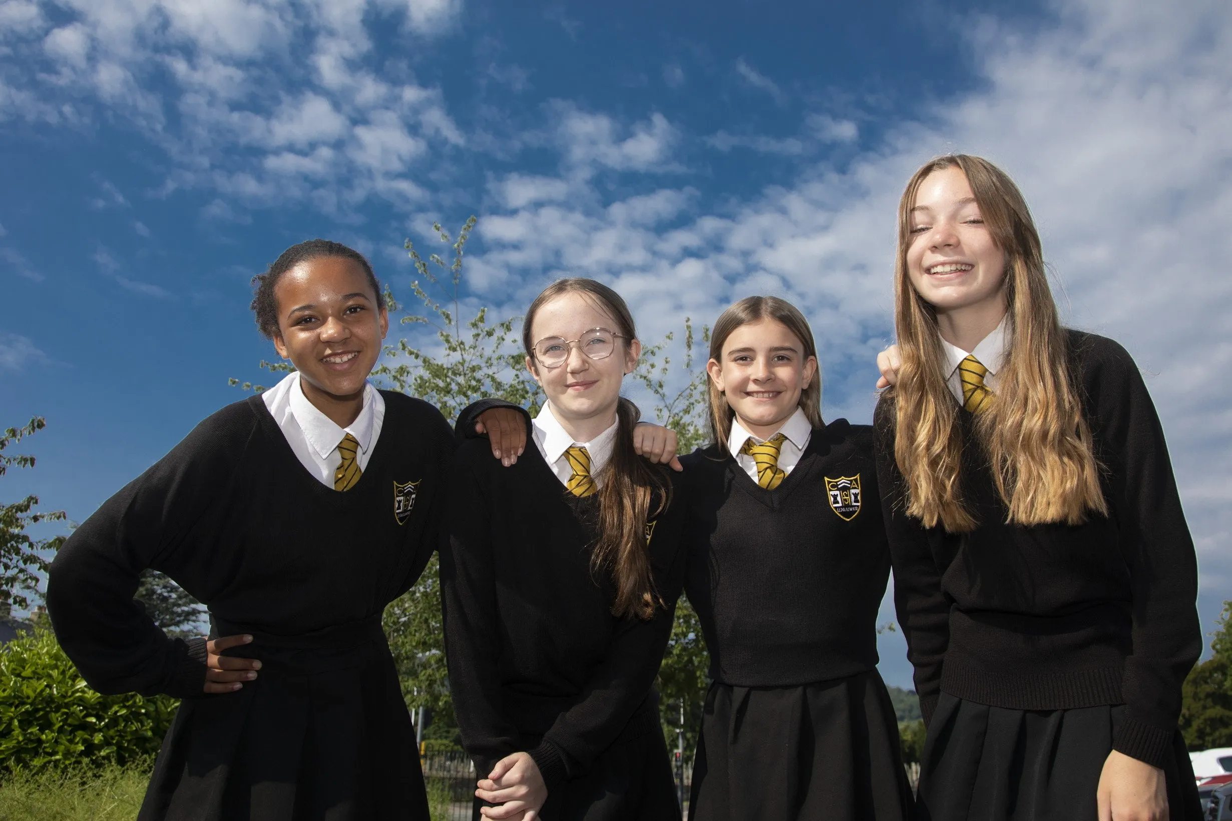 Four students in Castell Alun uniform standing together outside, smiling beneath a bright blue sky.