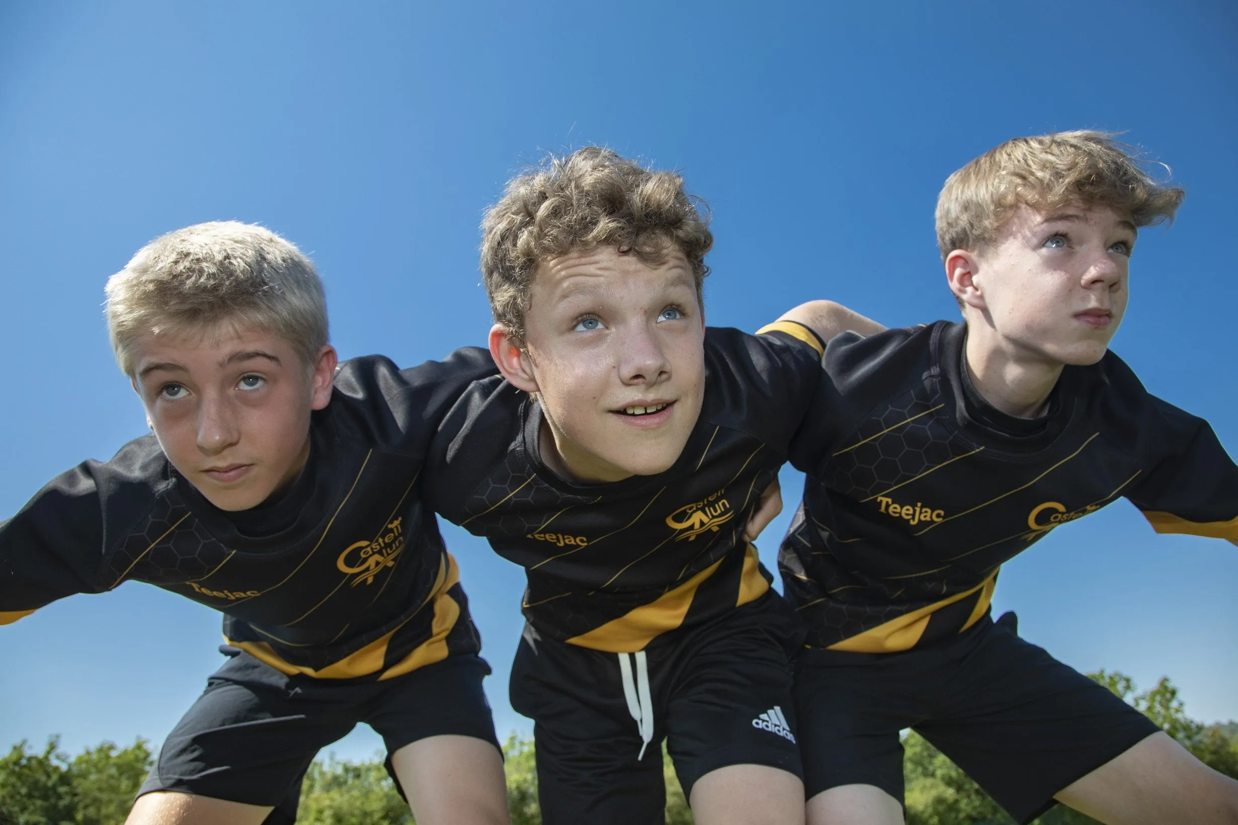 Three students in black and yellow sports kits crouched together on a sports field, posing for a team photograph against a clear blue sky.