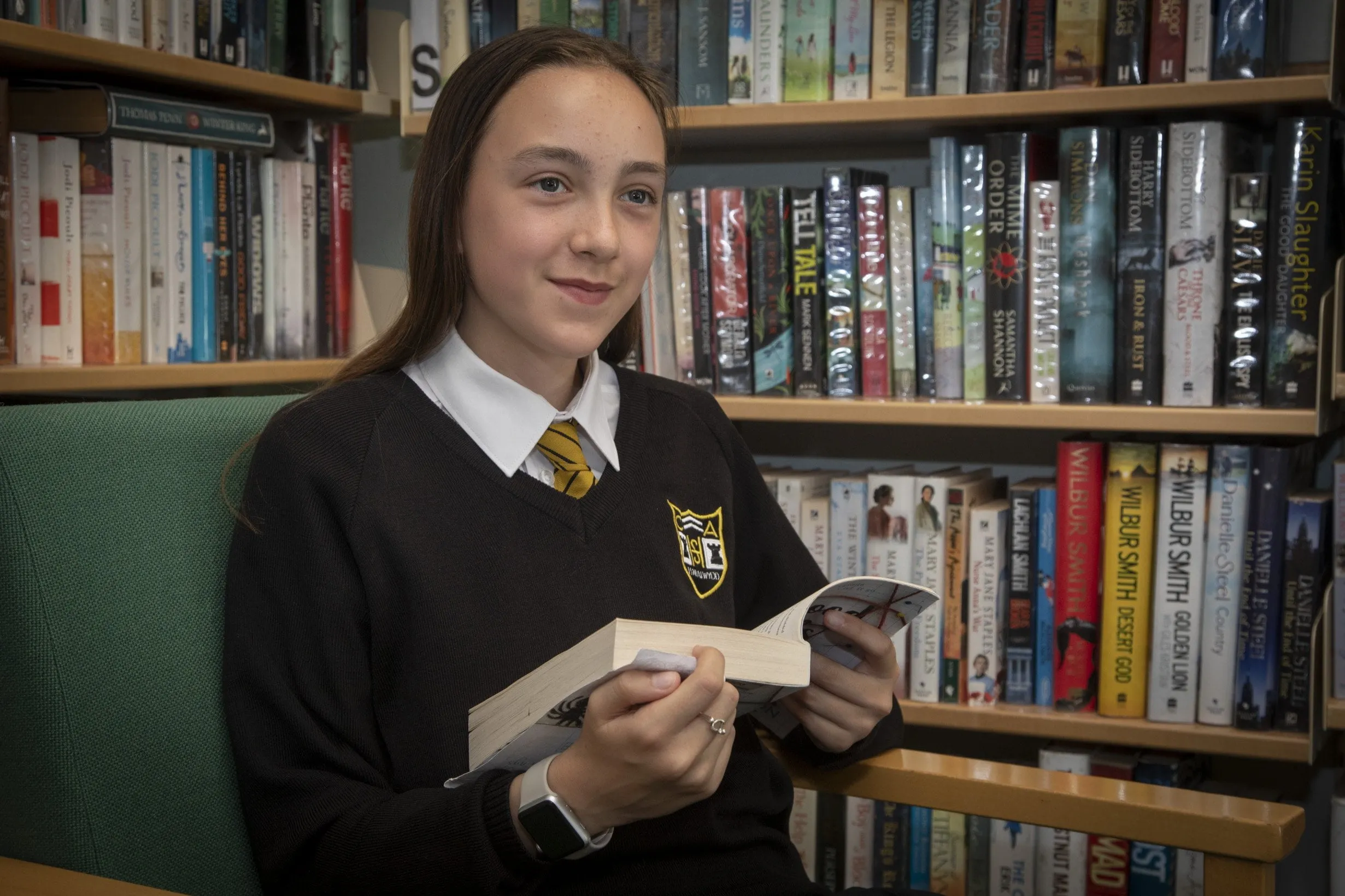 A student in school uniform sitting in a library chair and holding an open book, with shelves of books visible in the background.