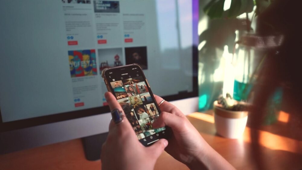 A person holds a smartphone showing a grid of social media photos while sitting at a desk in front of a computer monitor; soft daylight and a small potted plant are visible in the background.